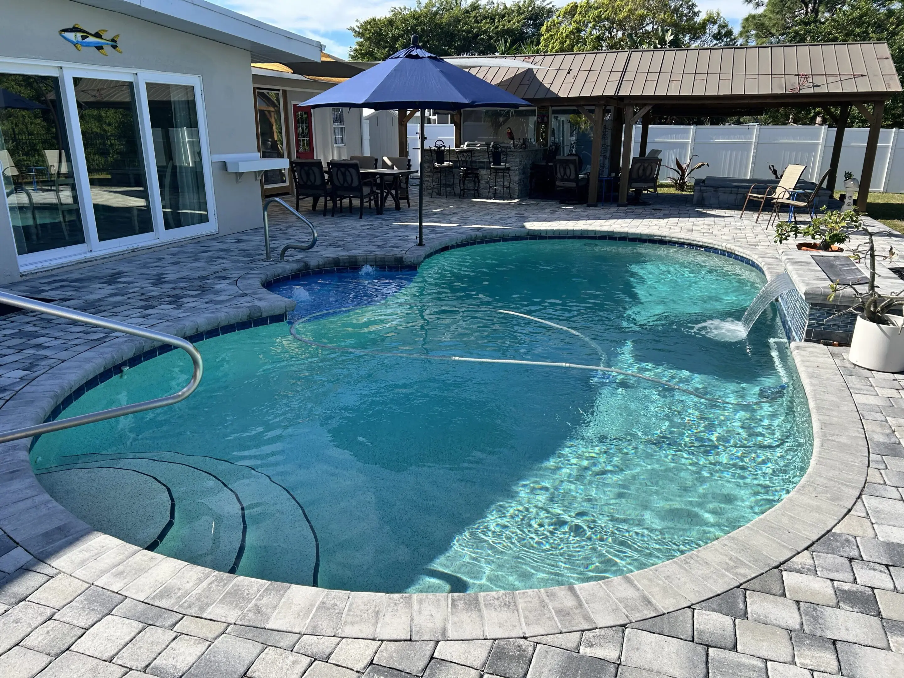 Clear backyard pool with stone deck and umbrella-shaded seating area.